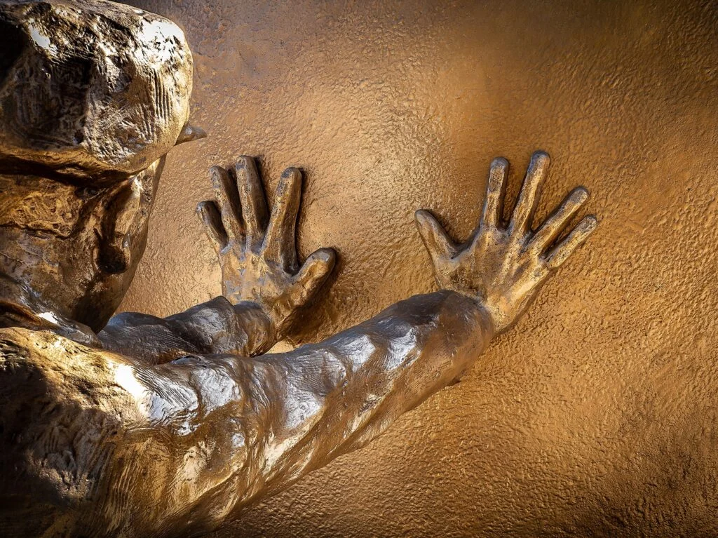 Manos de bronce apoyadas en un muro, con el metal brillante donde más se toca